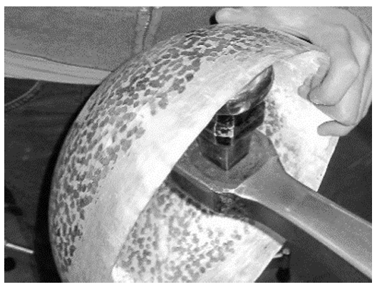 Hand hält gefleckten, runden Metallkörper; Schraubenschlüssel ist an einer Schraube angesetzt.